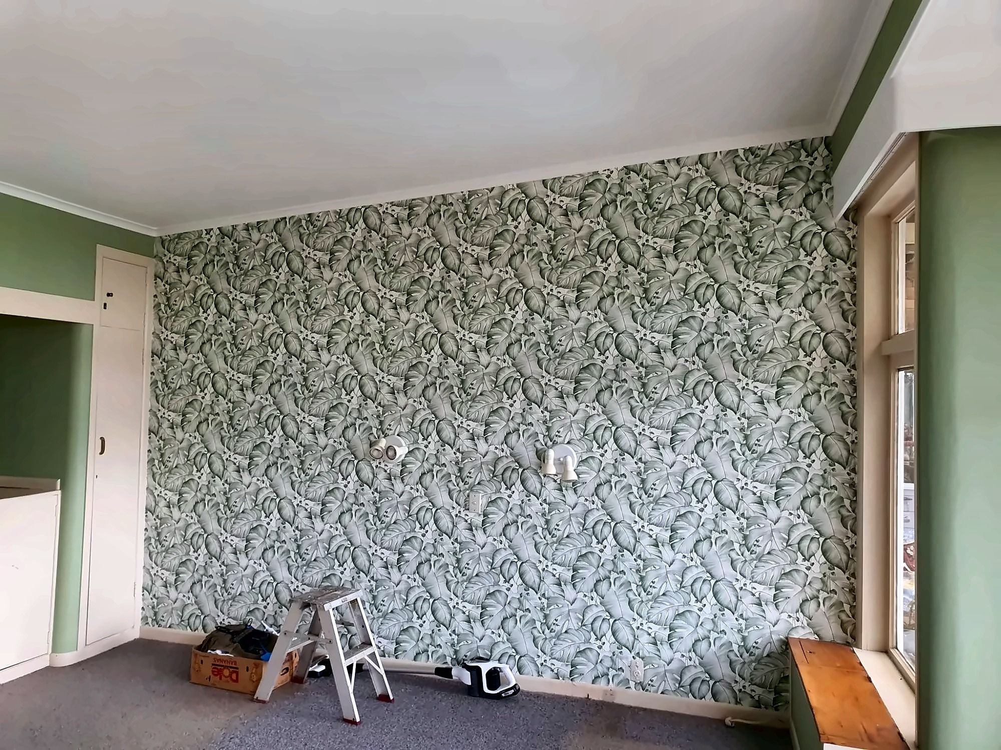 A full-height feature wall papered in a green and white leafy botanical print, in a room with sage-green walls.